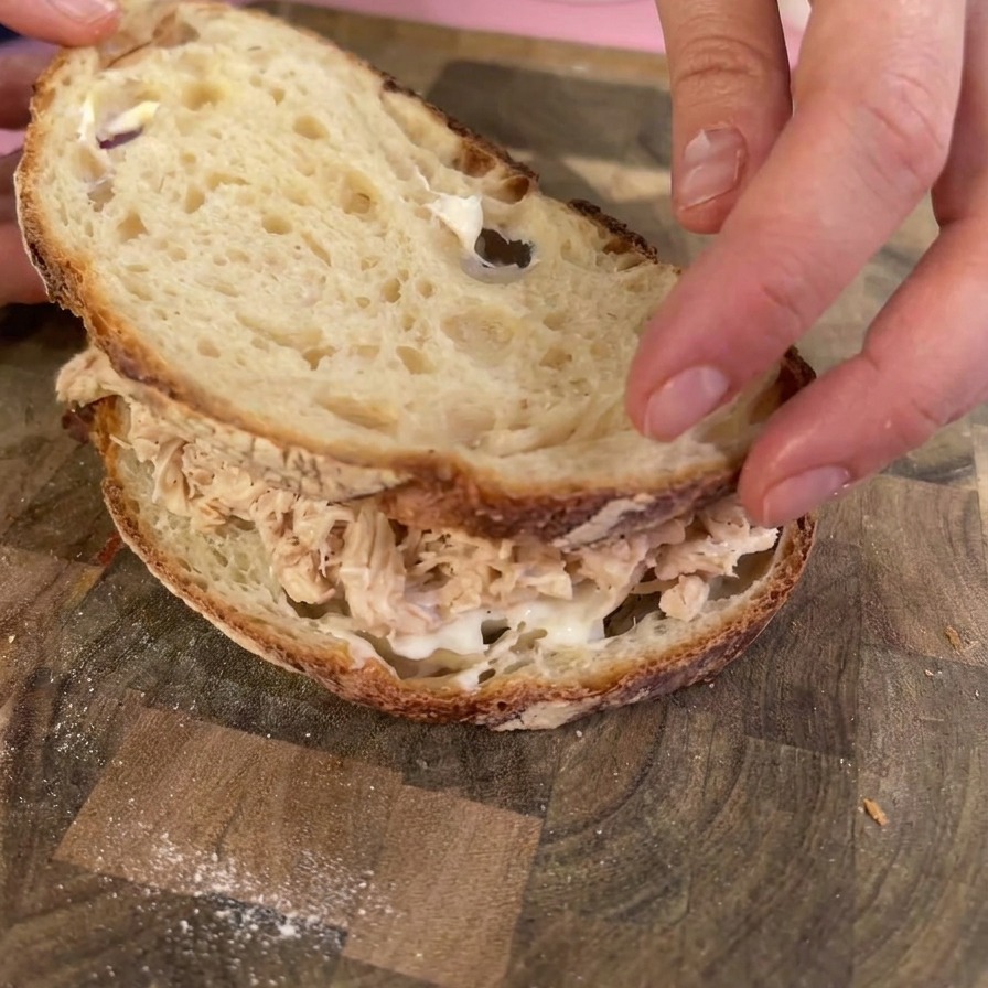 Manos colocando una rebanada tostada de pan de masa madre encima de una montaña de pollo desmechado para cerrar el sándwich.