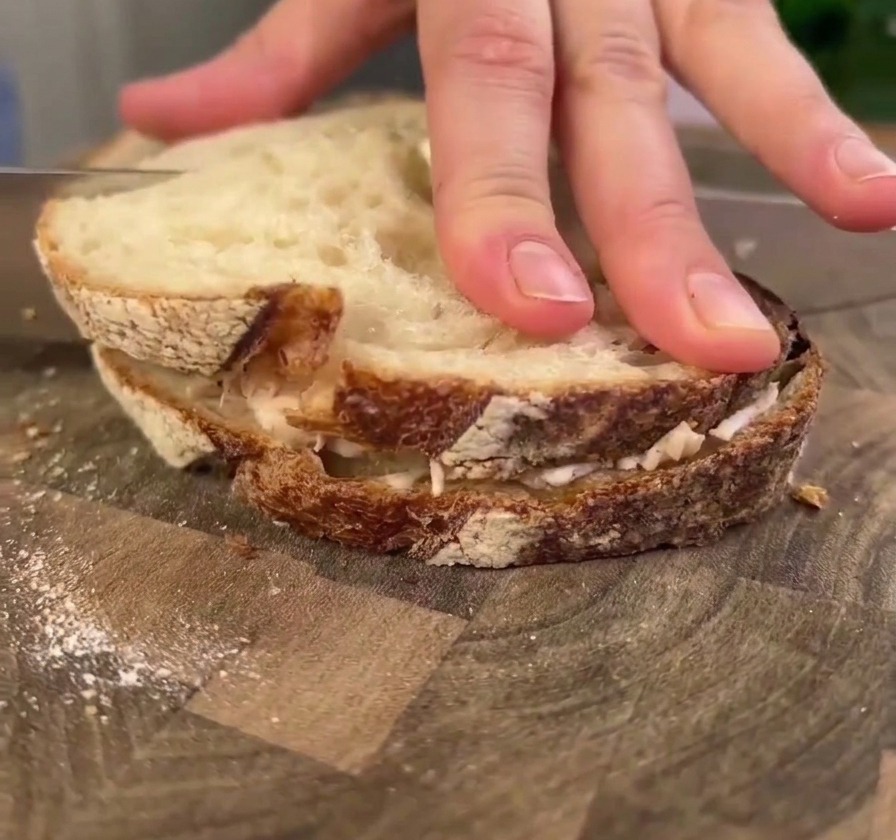 Una mano presionando firmemente hacia abajo sobre un sándwich grande de pollo y masa madre en una tabla de madera mientras un cuchillo se prepara para cortarlo.