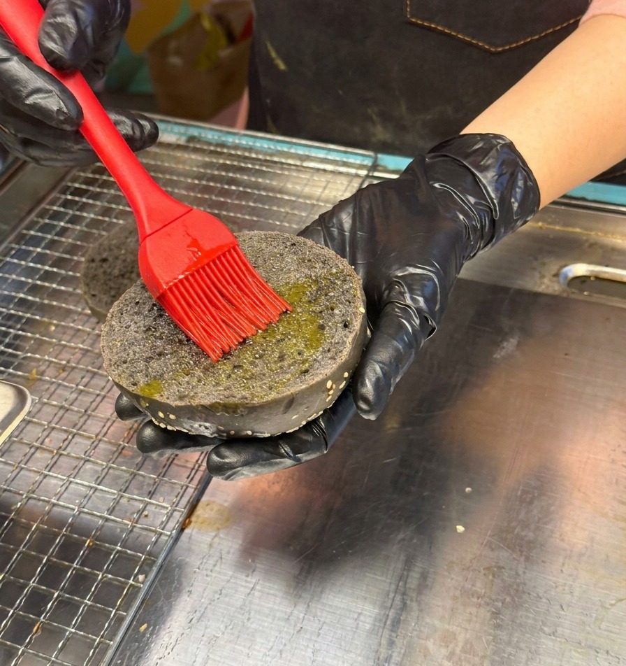 Mano con guante negro usando un pincel rojo de silicona para aplicar aceite en la cara interna de un pan negro con sésamo.