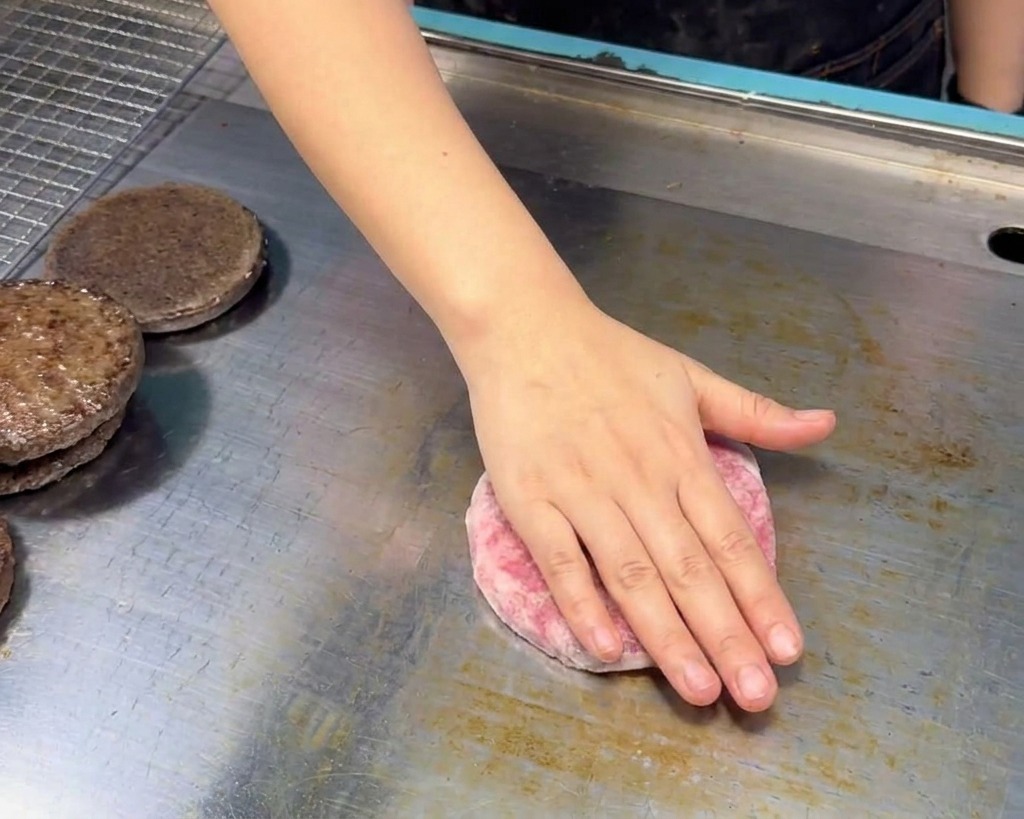 Mano colocando una hamburguesa de ternera cruda sobre una plancha de acero inoxidable junto a mitades de pan tostado.