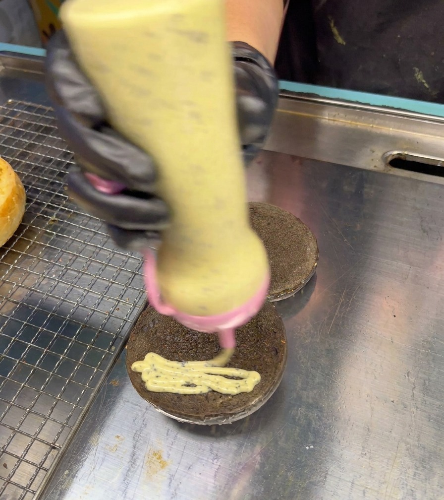 Biberón dispensador aplicando salsa cremosa de trufa en forma de zigzag sobre un pan negro de hamburguesa.