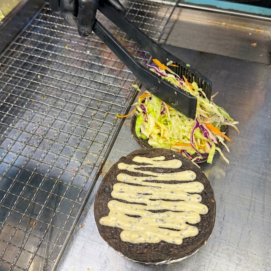 Persona usando unas pinzas para colocar una rodaja de tomate rojo brillante sobre la cama de verduras del pan al carbón.