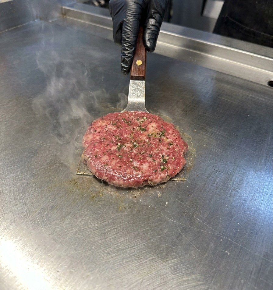 Espátula de metal presionando la carne de hamburguesa sobre una plancha de acero inoxidable con vapor a su alrededor.