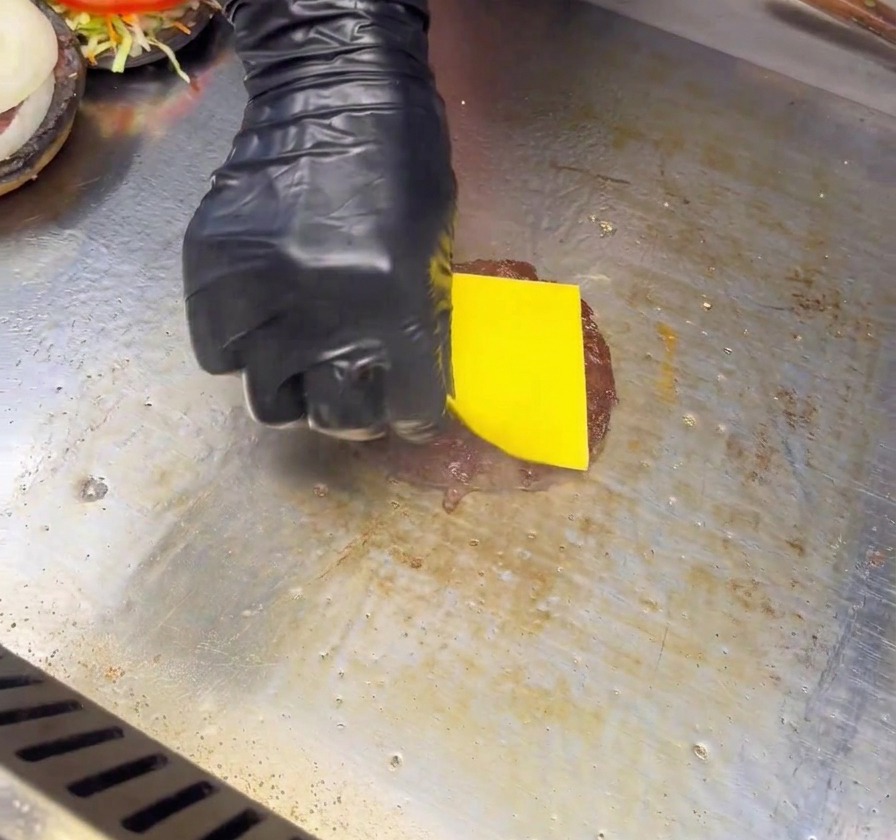 Mano con guante negro poniendo una loncha de queso amarillo sobre la carne en la plancha.