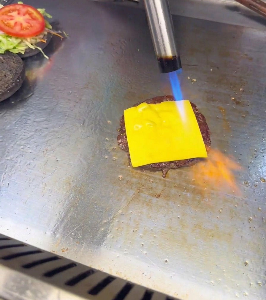 Llama azul y naranja de un soplete de cocina fundiendo una loncha de queso sobre una hamburguesa.