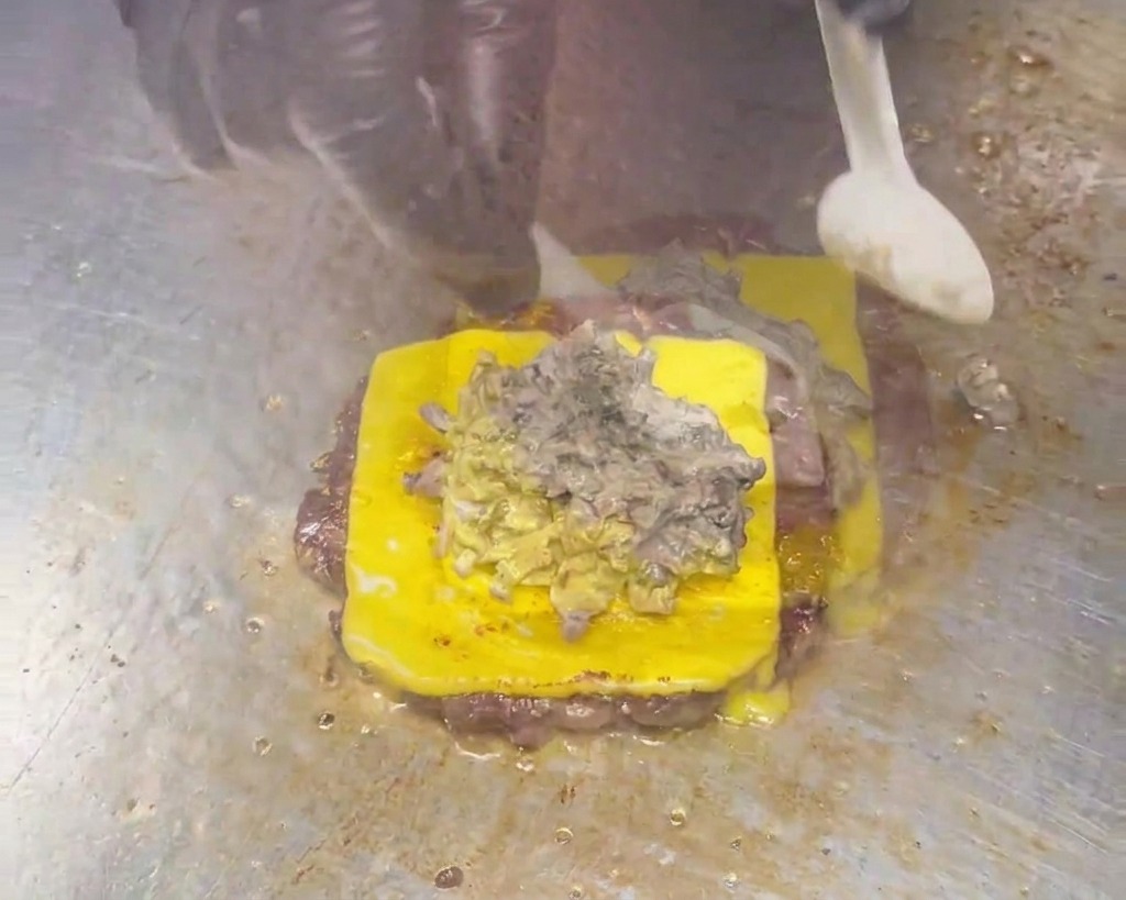 Cuchara esparciendo una salsa cremosa de champiñones color marrón grisáceo sobre una hamburguesa con queso.