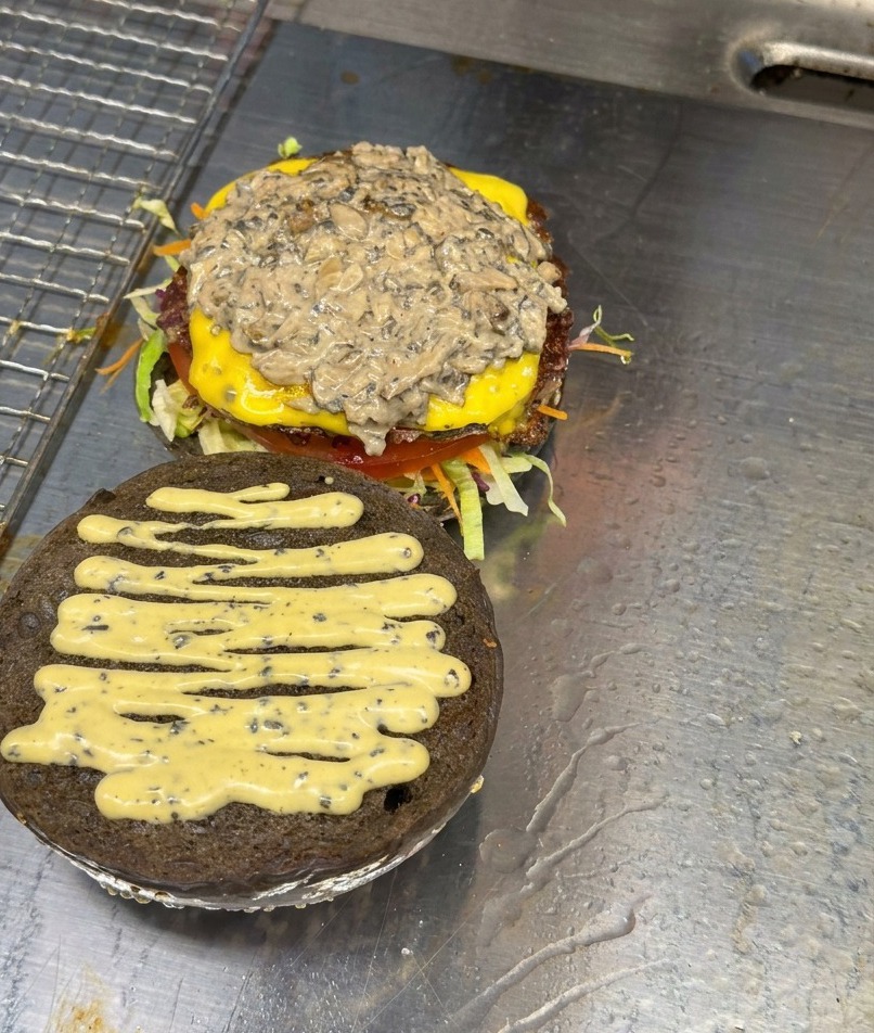 Pila de champiñones laminados salteados reposando sobre una hamburguesa montada con queso y salsa.
