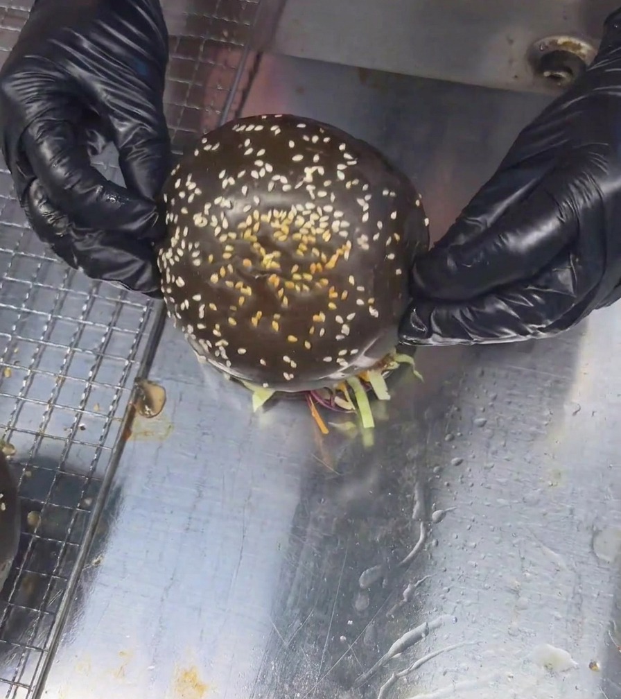Dos manos con guantes colocando un pan negro con sésamo sobre una hamburguesa de ternera y champiñones totalmente montada.