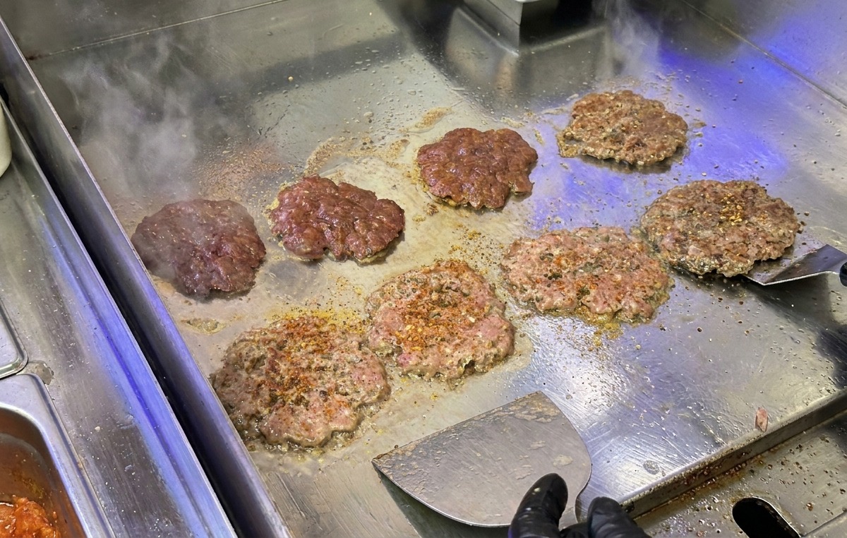 Carnes de hamburguesa siendo aplastadas y selladas en una plancha de acero inoxidable con una espátula metálica de cocina.