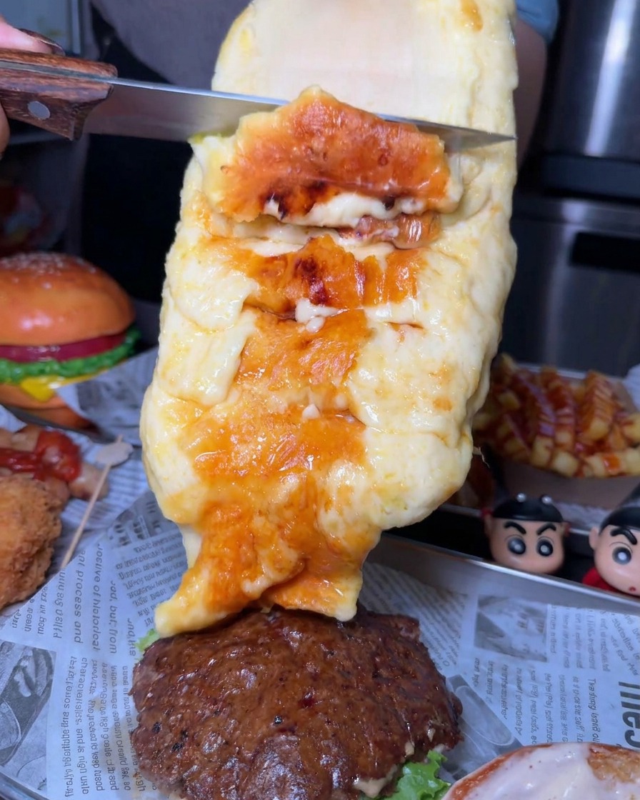 Una capa generosa de queso fundido y dorado siendo raspada de una gran rueda con un cuchillo hacia una hamburguesa.