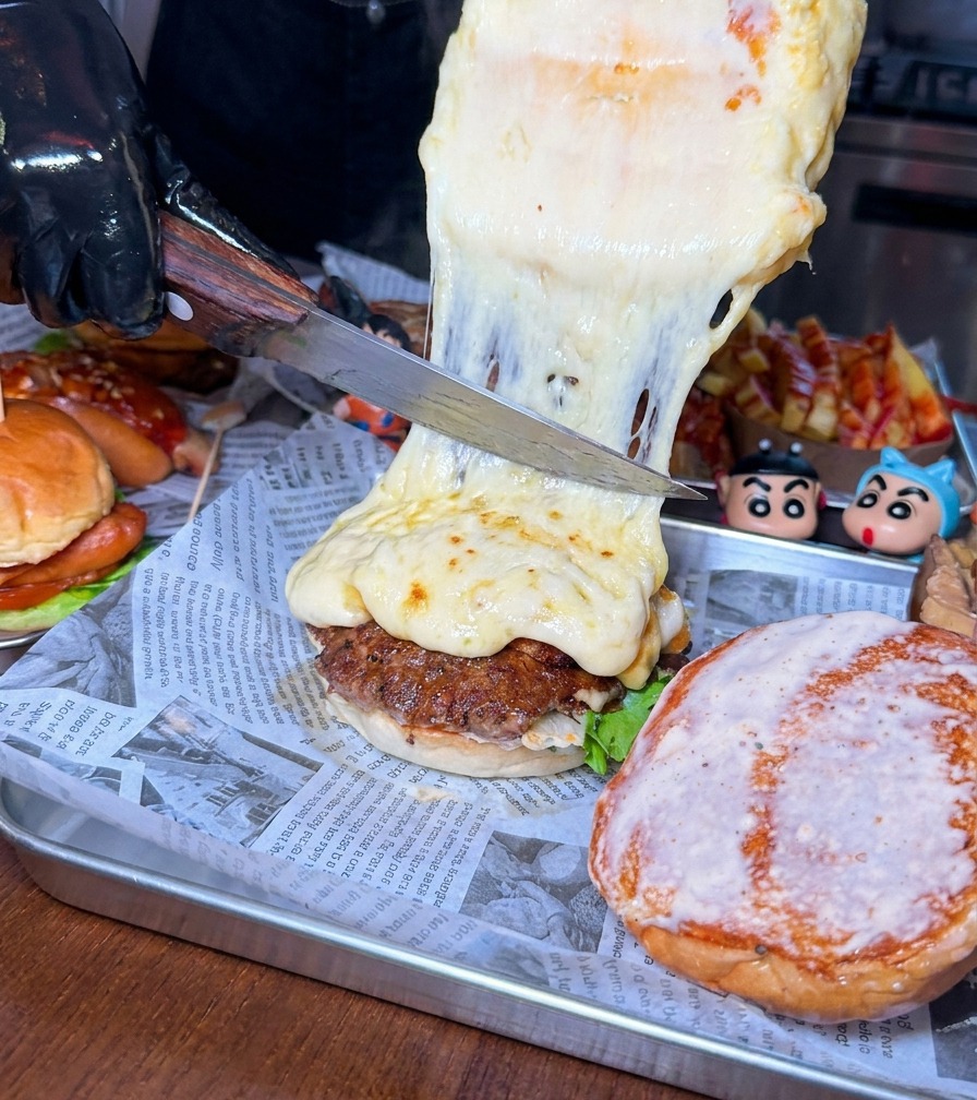 Un cuchillo grande raspando una capa gruesa de queso fundido y burbujeante desde la rueda hacia una hamburguesa.