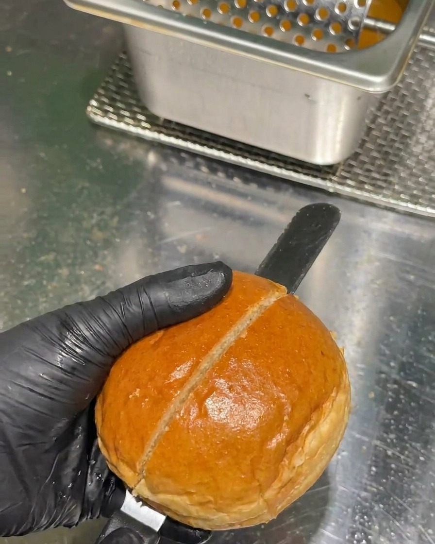 Una mano con guante negro usa un cuchillo de sierra largo para cortar un pan de hamburguesa dorado por la mitad en una encimera de acero inoxidable.