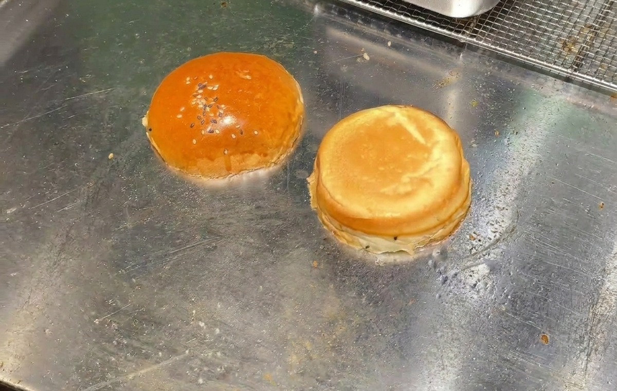 Dos mitades de un pan de hamburguesa tostándose boca abajo sobre la superficie de una plancha metálica plana.