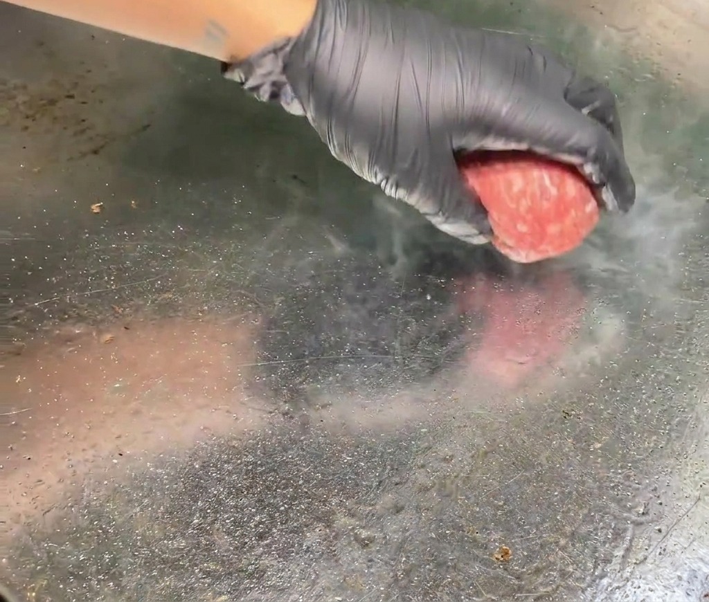 Una mano con guante coloca una bola de carne de res molida cruda sobre una plancha de metal caliente con vapor saliendo del punto de contacto.