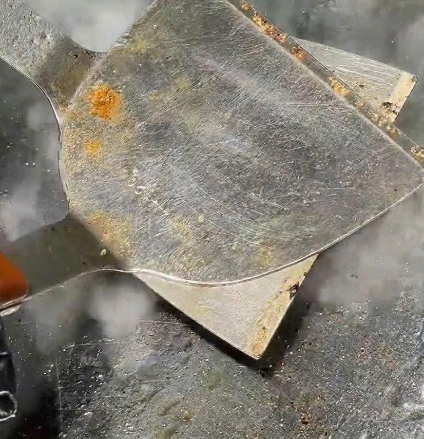 Una espátula de metal aplasta una bola de carne hasta formar una hamburguesa delgada en una plancha caliente con vapor subiendo.