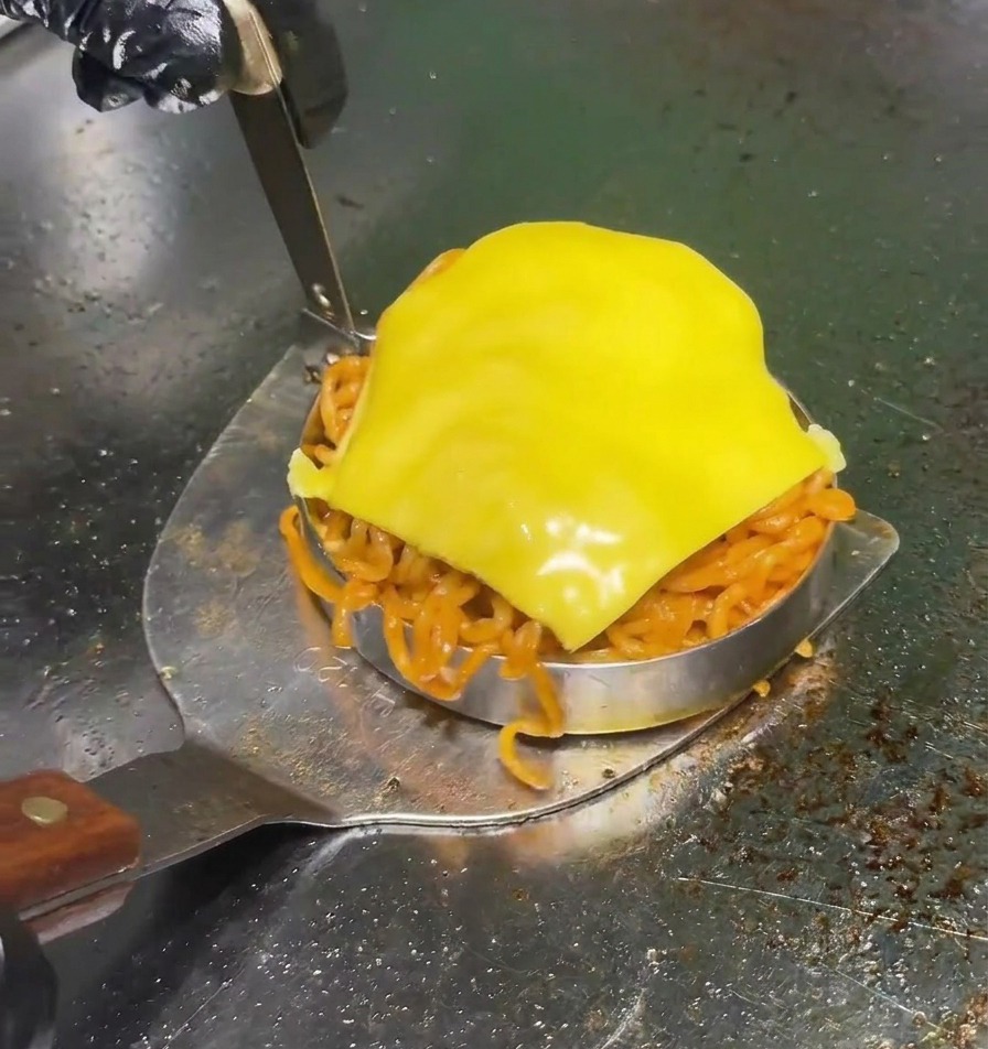 Una espátula de metal levanta un molde circular lleno de fideos rojos picantes y coronado con una rebanada de queso amarillo en una plancha profesional.