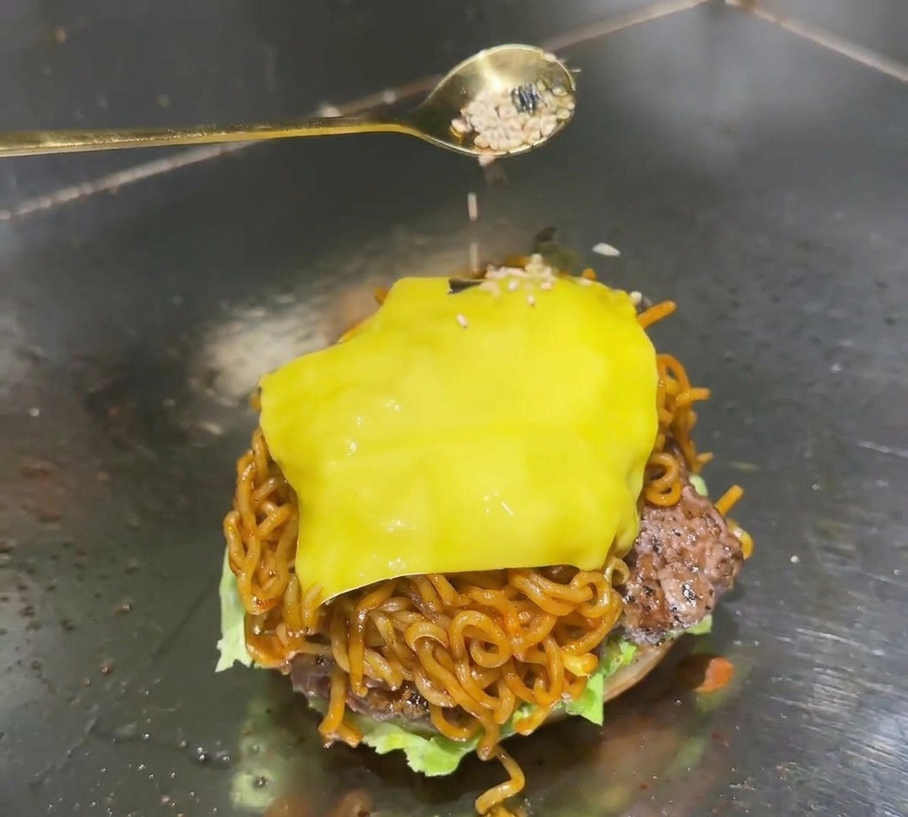 Una cuchara dorada espolvorea una mezcla de semillas de sésamo blanco y copos de alga negra sobre el queso derretido de una hamburguesa de ramen.