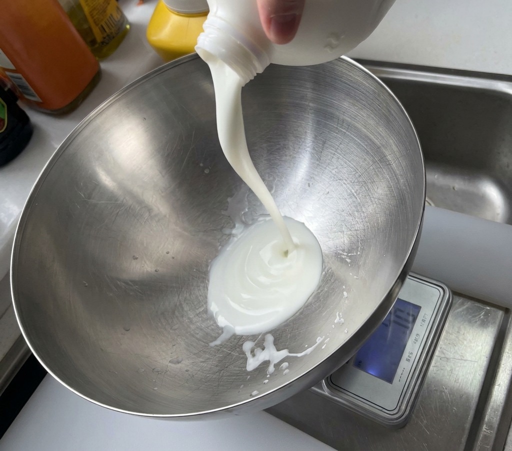 Un chorro de yogur blanco vertiéndose desde un envase plástico a un tazón grande de metal cepillado.