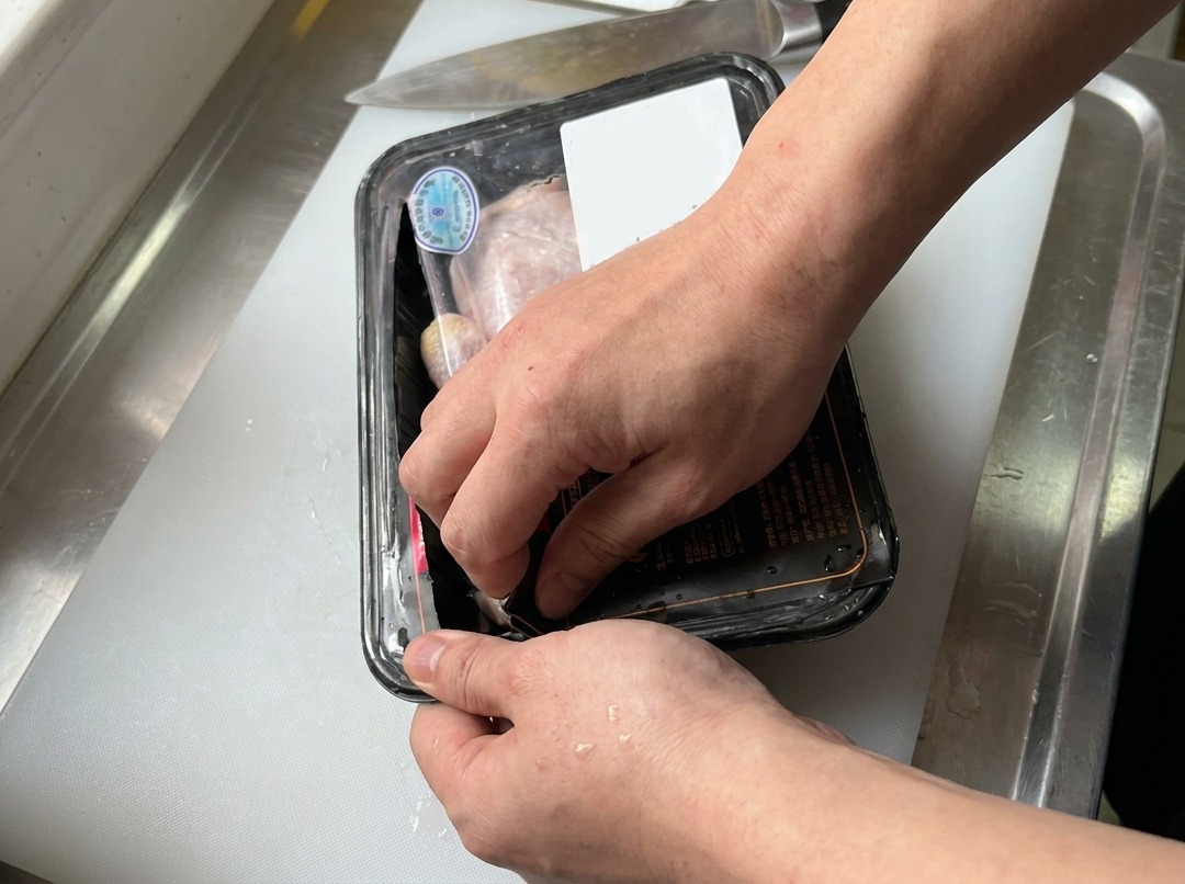 Manos abriendo una bandeja envuelta en plástico con contramuslos de pollo crudos sobre una tabla de cortar blanca junto a un cuchillo de cocina.