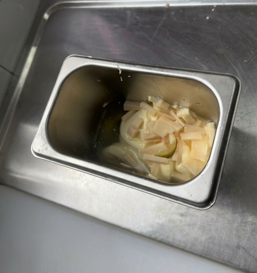 Un recipiente de acero inoxidable con una cucharada de mayonesa, mostaza amarilla y trocitos de pepinillos verdes picados.