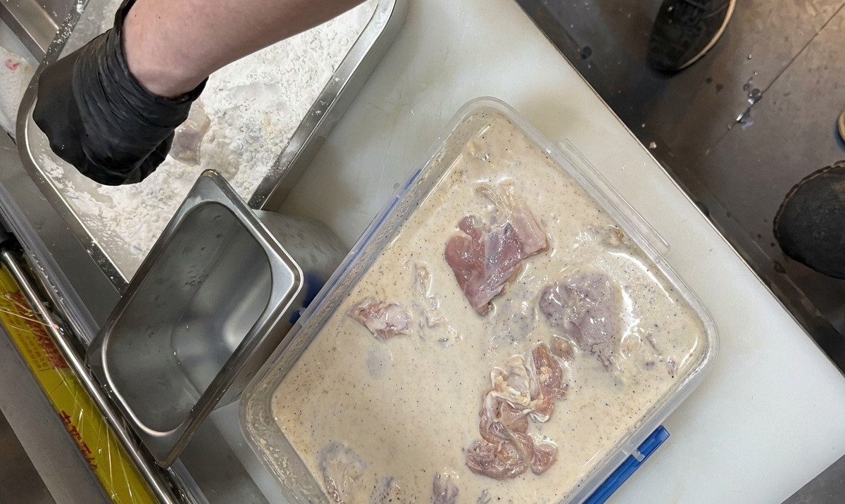 Manos con guantes pasando pollo marinado en un líquido lechoso a una bandeja de harina de rebozado sazonada.