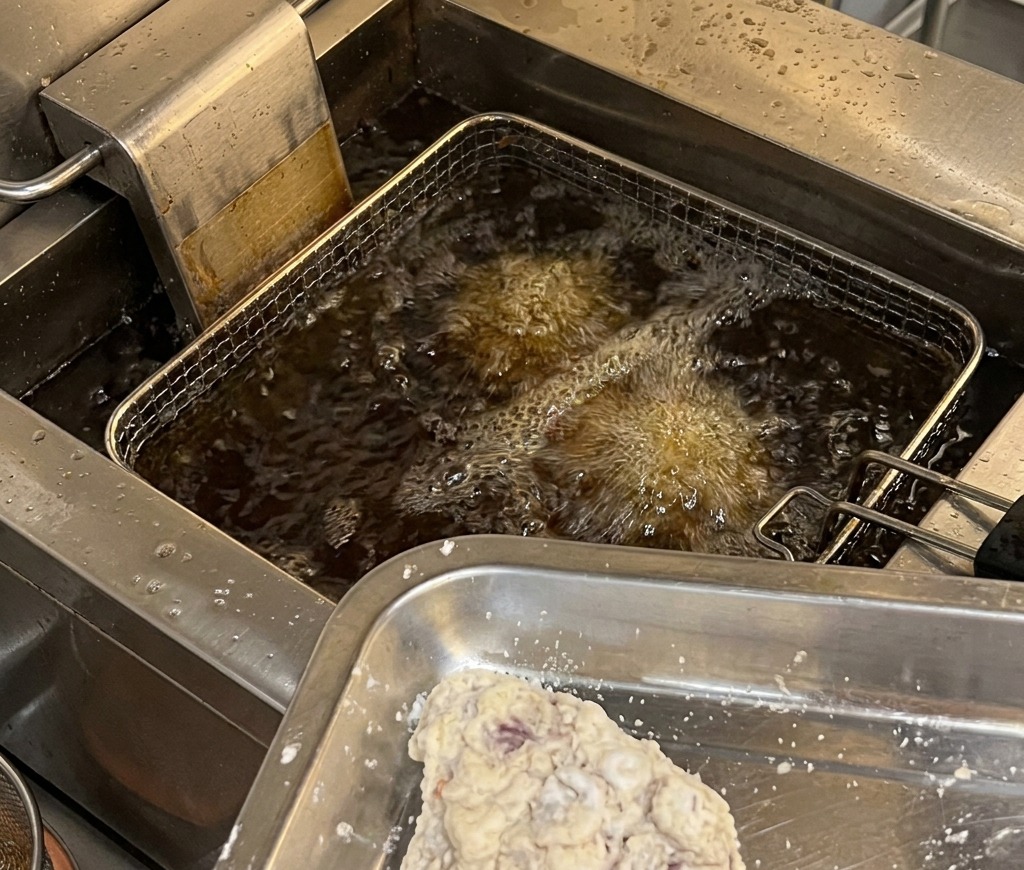 Trozos de pollo rebozados en una bandeja de metal listos para entrar en una freidora industrial con aceite caliente y burbujeante.