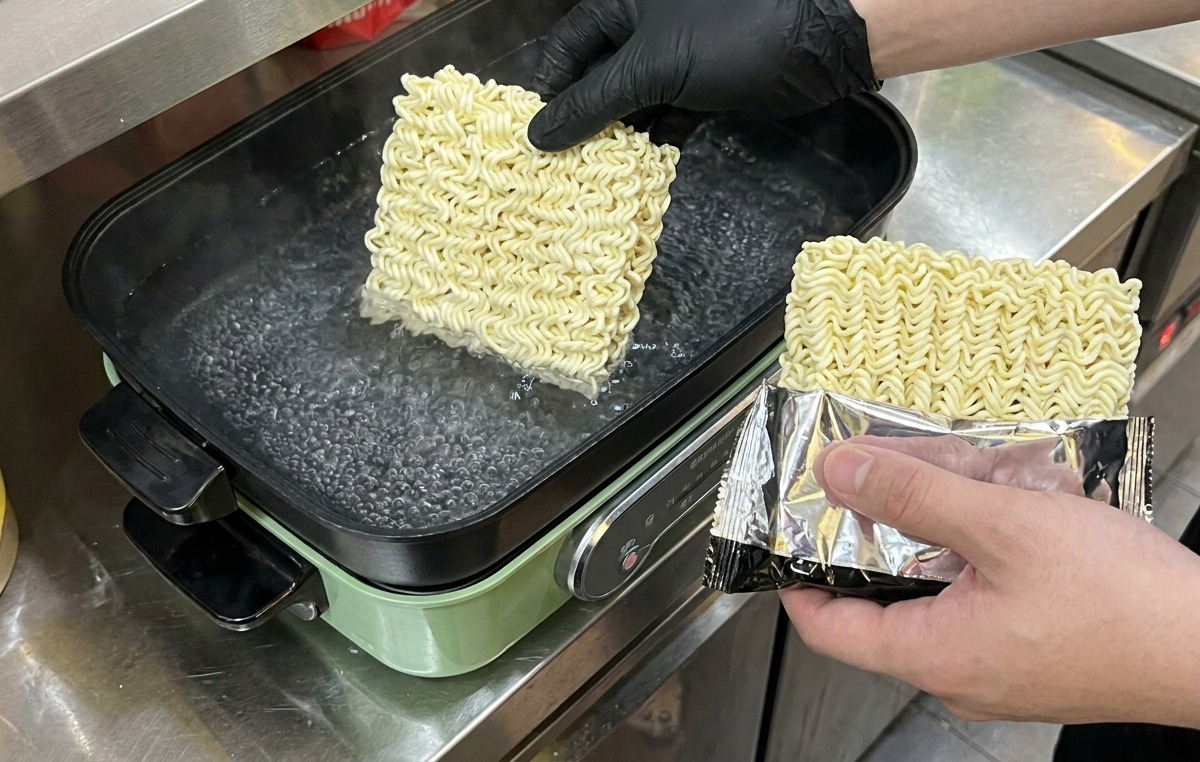 Una persona con guantes negros dejando caer bloques cuadrados de fideos instantáneos secos en una olla de agua hirviendo.