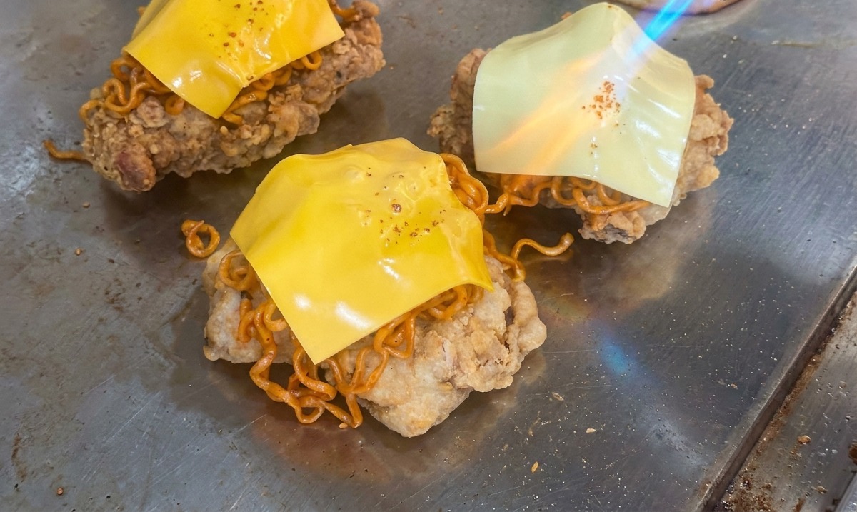 La llama de un soplete derritiendo un cuadrado de queso amarillo sobre una pila de fideos picantes y pollo frito en una parrilla.