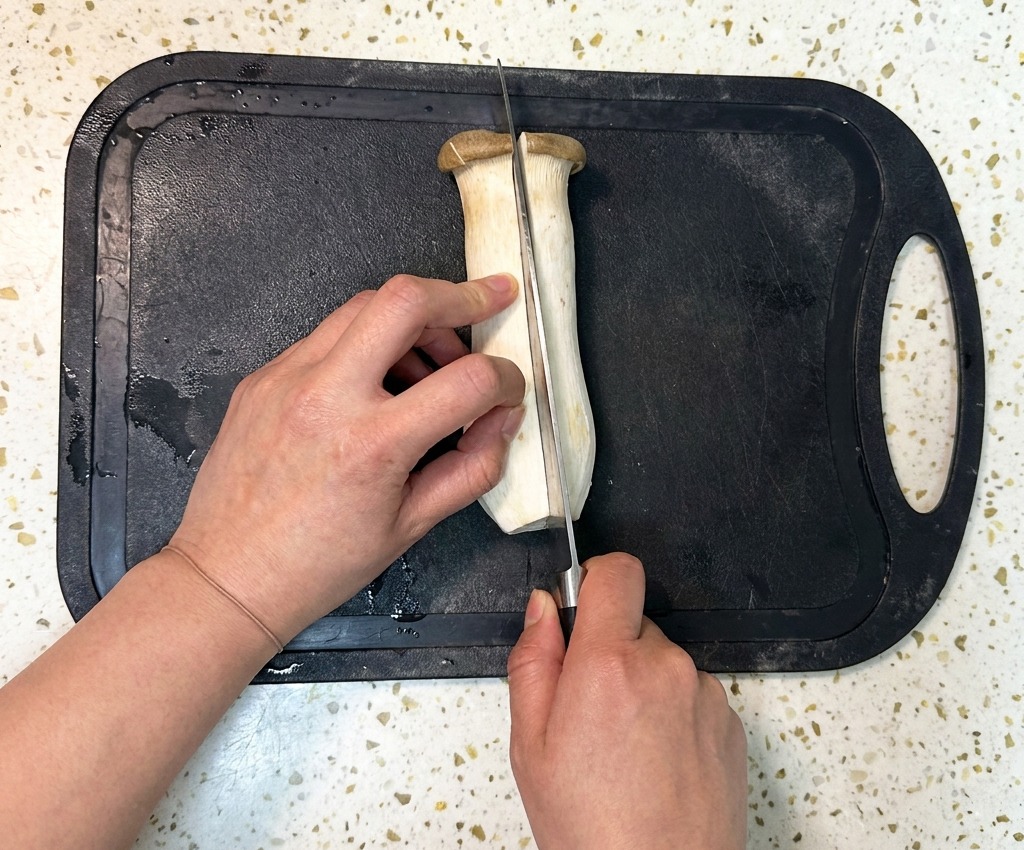 Una persona usando un cuchillo de chef para cortar una gran seta eryngii por la mitad a lo largo sobre una tabla de picar de plástico negro.