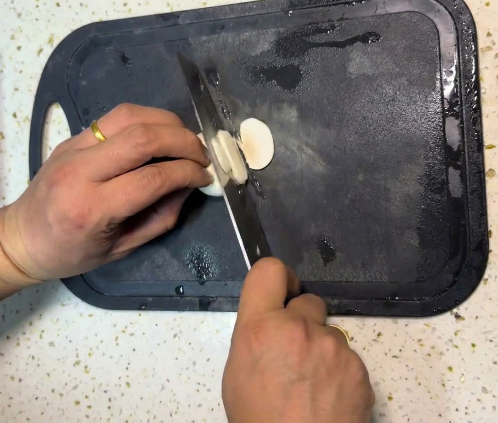 Las manos de una persona usando un cuchillo de chef para cortar champiñones blancos en rodajas finas sobre una tabla de picar negra.