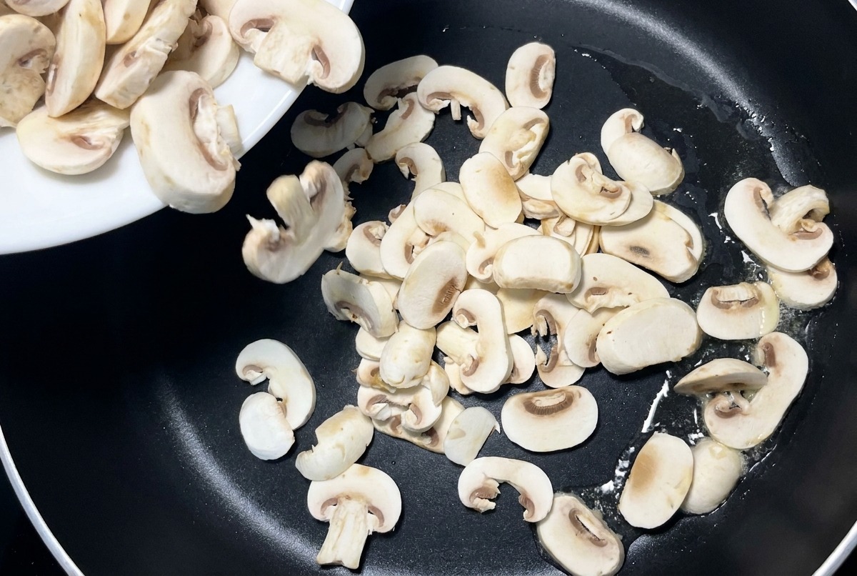 Champiñones laminados cayendo desde un plato de cerámica blanca hacia una sartén antiadherente negra.