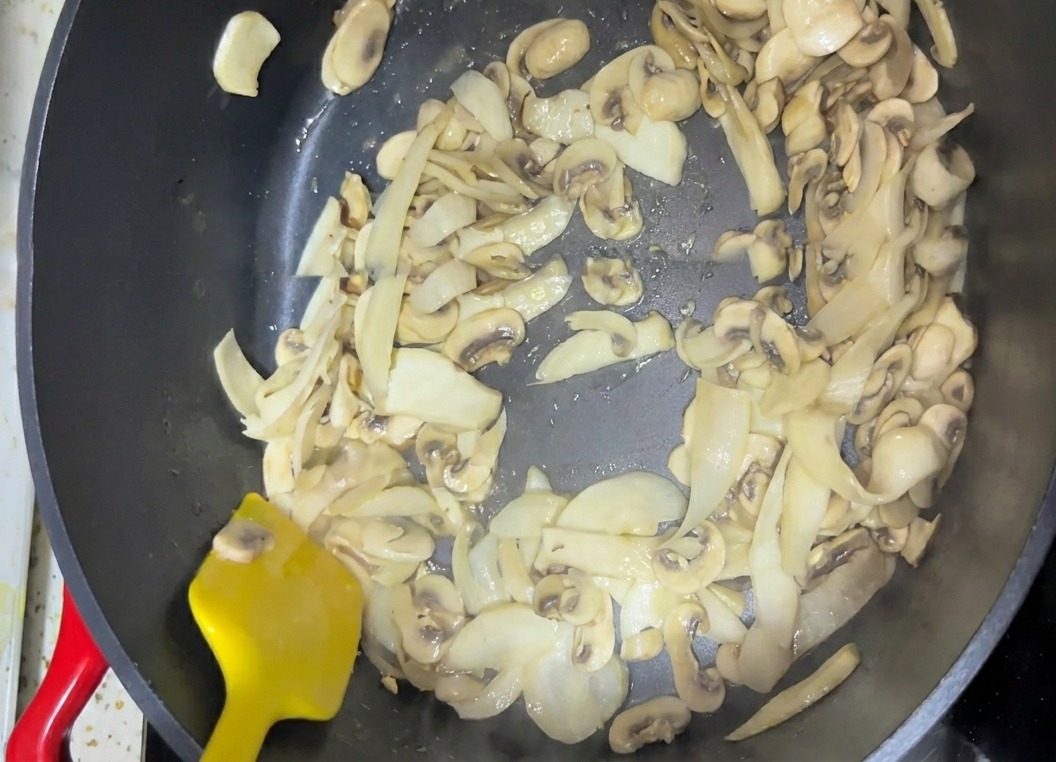Champiñones laminados siendo removidos en una sartén oscura con una espátula de color amarillo brillante.