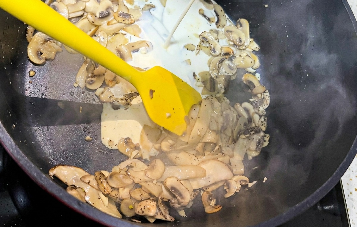 Un chorro de crema de leche blanca siendo vertido en una sartén negra llena de champiñones salteados.