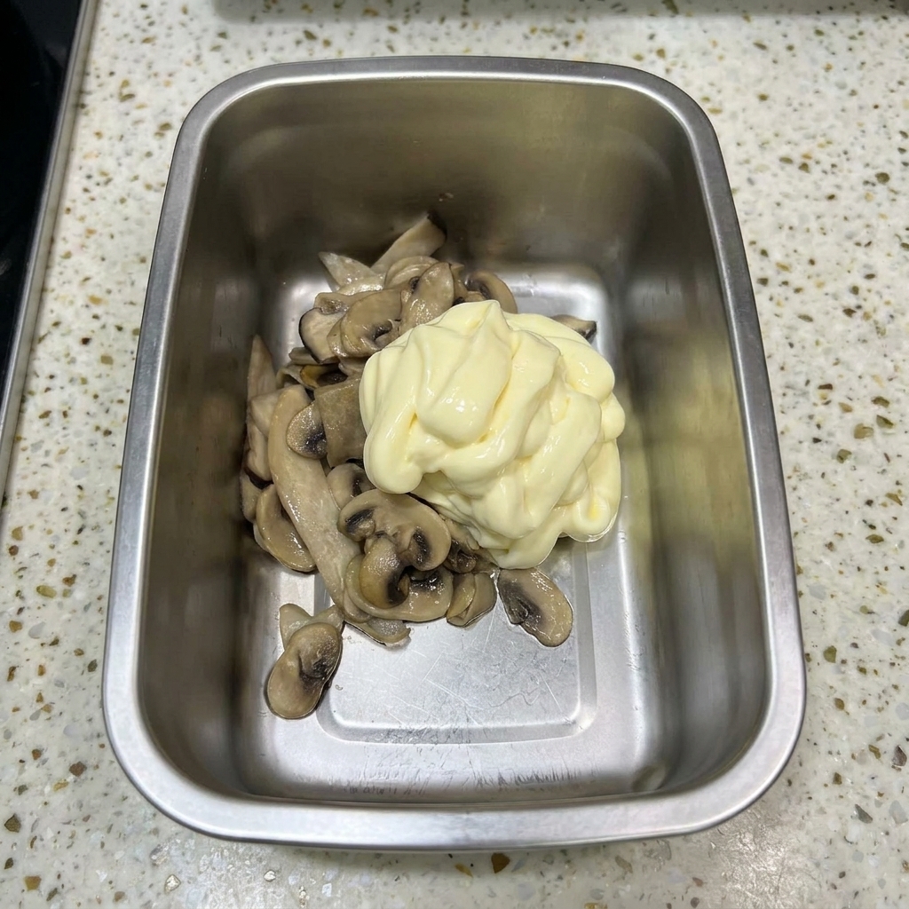 Un recipiente de metal con champiñones salteados cubiertos por un gran copo de mayonesa de color amarillo pálido.