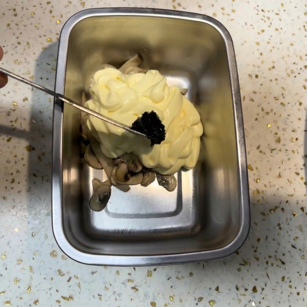 Un pequeño utensilio de metal colocando una porción de pasta de trufa negra oscura sobre un montículo de mayonesa y champiñones.