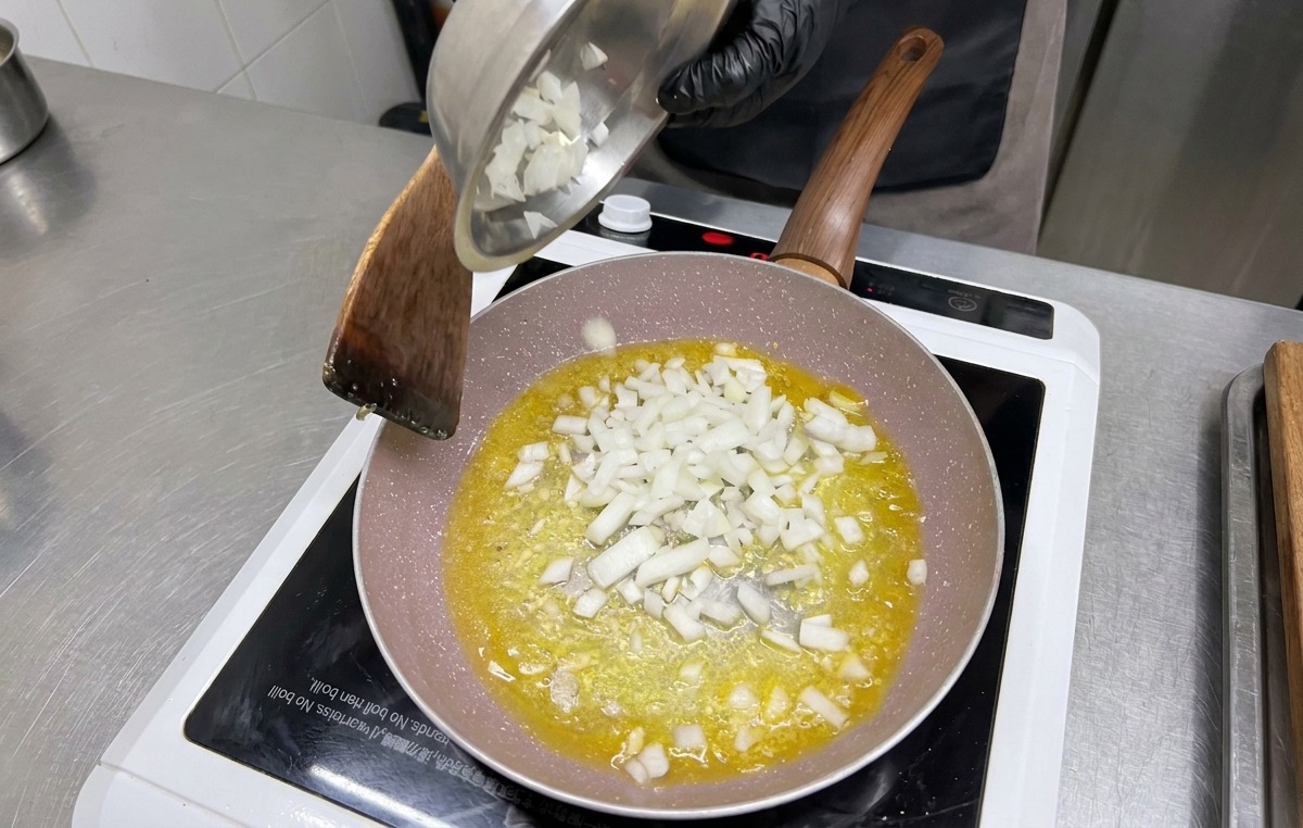 Cebollas blancas picadas en cubos vertiéndose desde un tazón de metal a la sartén sobre el ajo y la mantequilla.