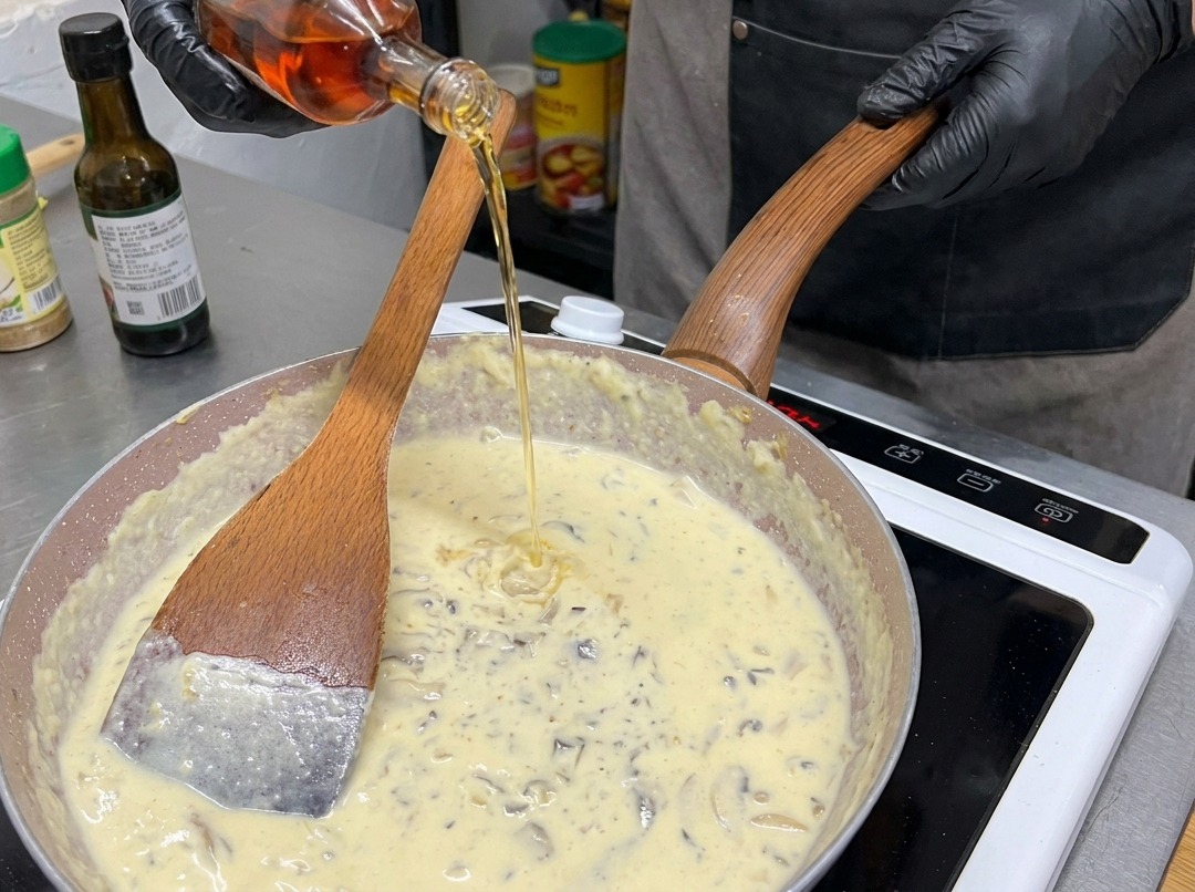 Una persona con guantes negros vertiendo un líquido ámbar desde una botella de vidrio en una sartén llena de salsa de champiñones cremosa.