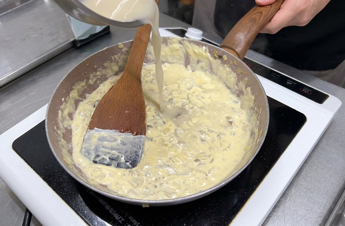 Caldo claro vertiéndose desde un tazón de acero inoxidable en una sartén de champiñones y crema hirviendo a fuego lento.