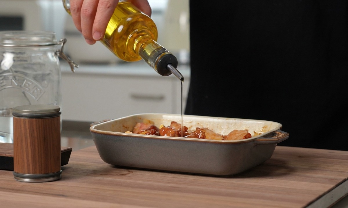 Aceite de cocina dorado vertiéndose desde una botella de vidrio sobre costillas de cerdo sazonadas en una fuente de cerámica gris para hornear.