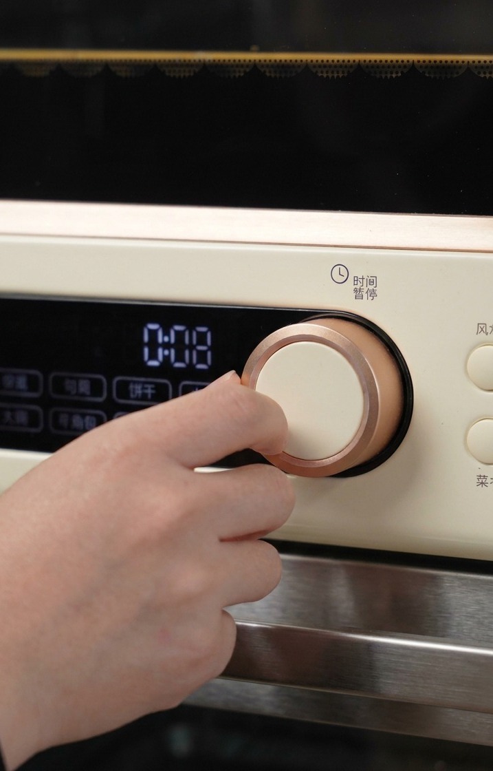 Una mano girando el dial de oro rosa de un horno de color crema para programar el tiempo y la temperatura iniciales de horneado.