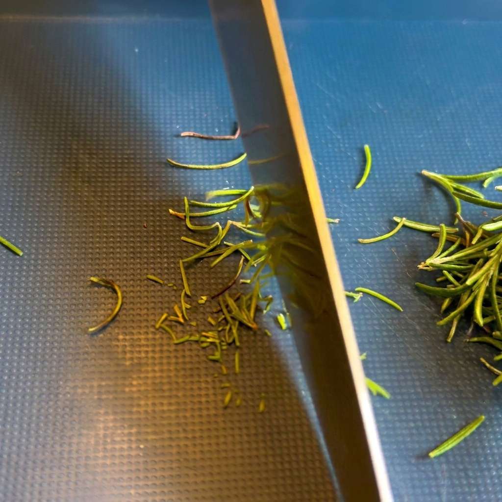 Primer plano de la hoja de un cuchillo de chef picando hojas verdes de romero sobre una tabla de cortar oscura y texturizada.