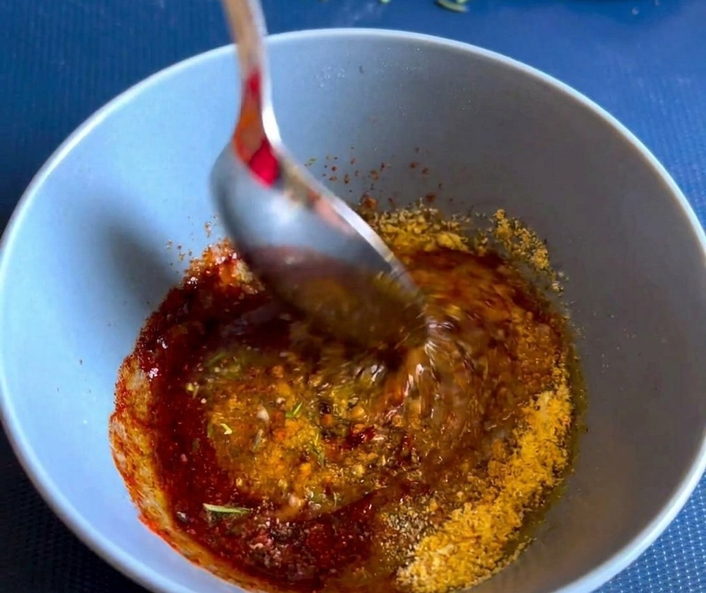Una cuchara de metal remueve una pasta espesa de especias y hierbas de color marrón rojizo dentro de un pequeño bol de cerámica azul.