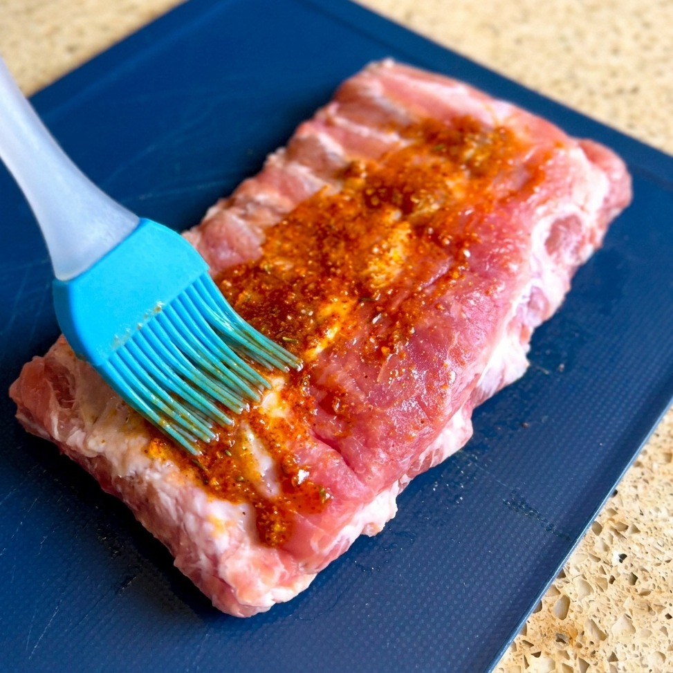 Un pincel de silicona azul extiende un marinado espeso y granuloso de color rojo sobre la superficie de un costillar de cerdo crudo en una tabla azul.