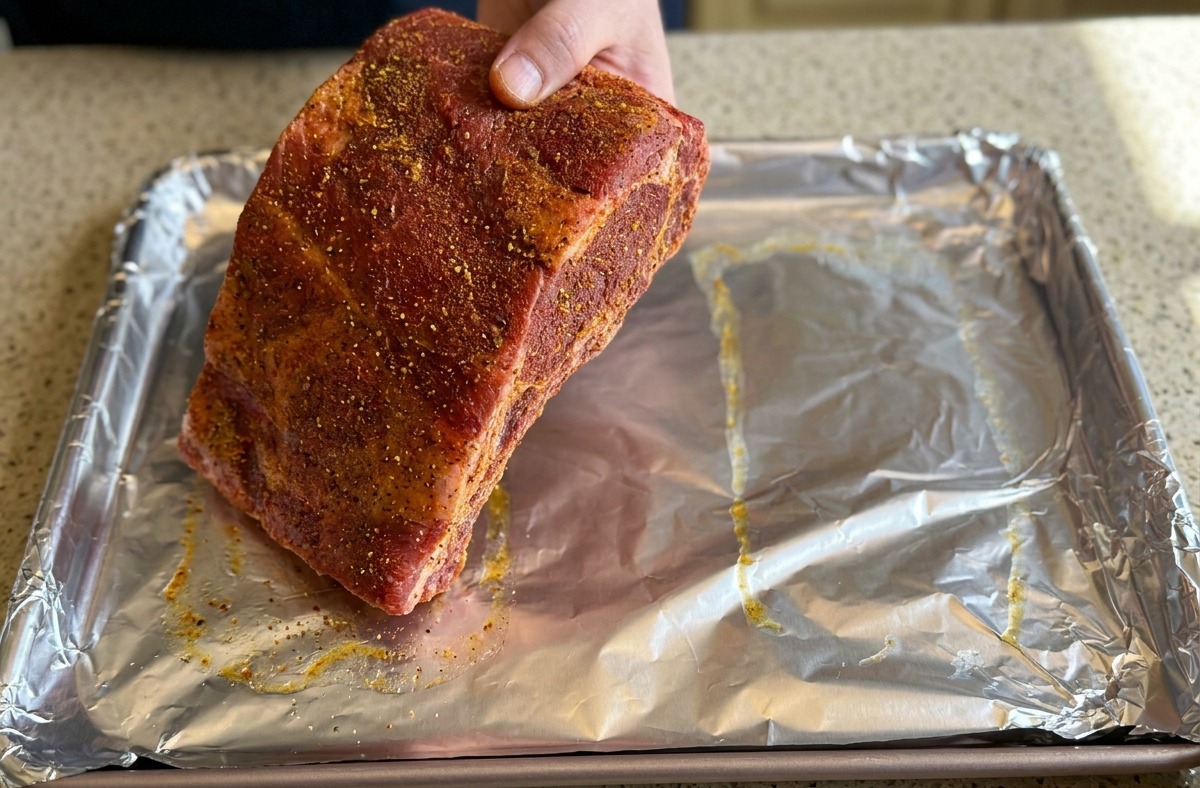 Una mano coloca un costillar de cerdo grande y sazonado sobre una bandeja de horno cubierta con papel de aluminio plateado arrugado.