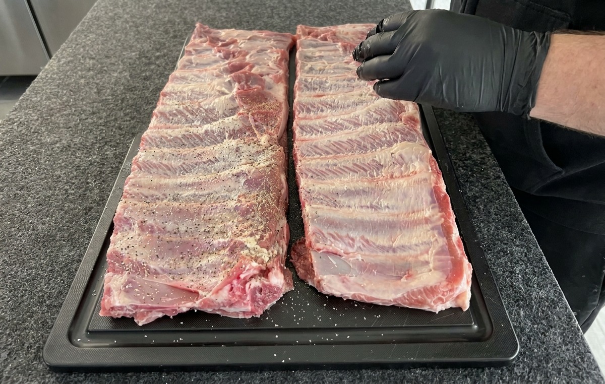 Dos costillares de cerdo crudos siendo sazonados con una mezcla de sal, pimienta y ajo en polvo sobre una tabla de cortar negra.
