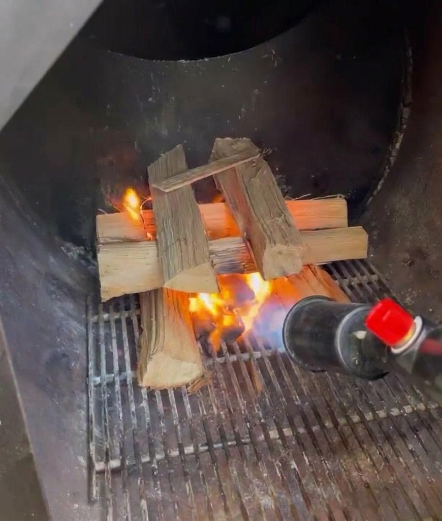 Un soplete de propano encendiendo una pila de leños de roble partidos dentro de la caja de fuego metálica de un ahumador.