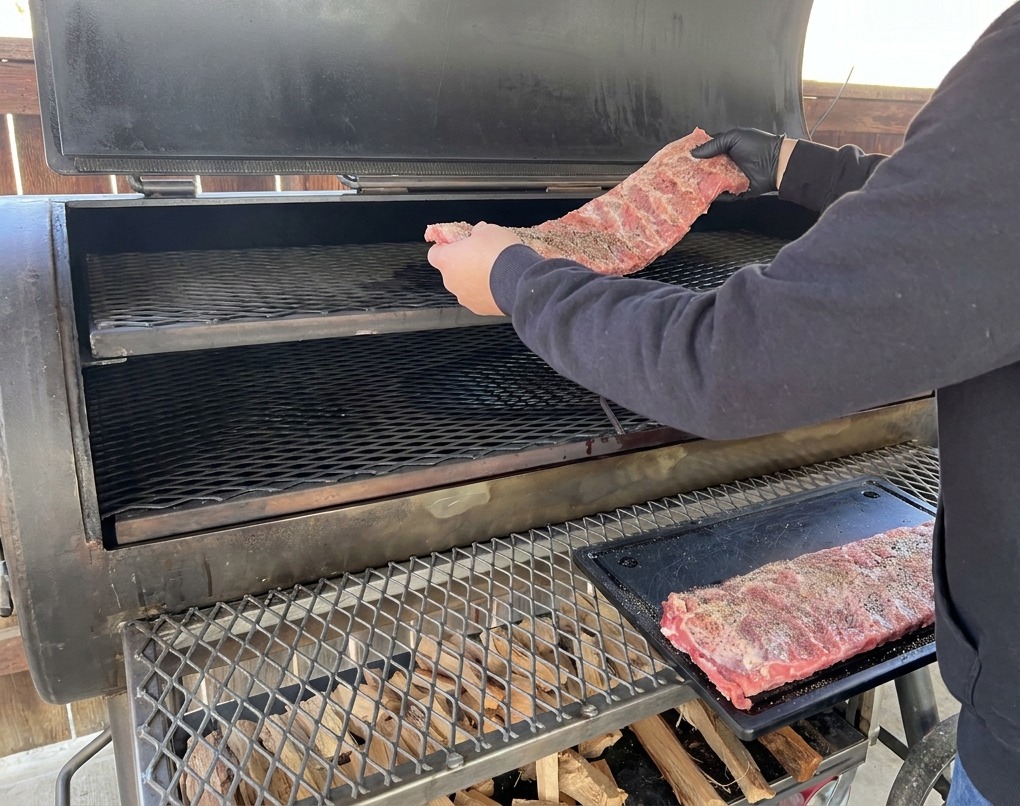 Una persona con sudadera negra y guantes negros colocando un costillar de cerdo sazonado en la rejilla superior de un gran ahumador de flujo desfasado.