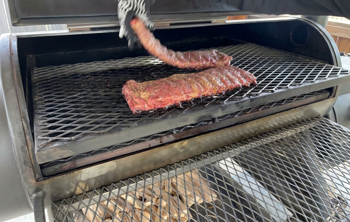 Una mano con un guante texturizado resistente al calor volteando un costillar de costillas parcialmente ahumadas en la rejilla metálica de un ahumador.