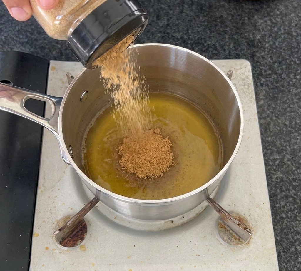 Una mano vertiendo azúcar moreno desde un frasco transparente en una olla de acero inoxidable que contiene vinagre de sidra de manzana sobre un quemador portátil.