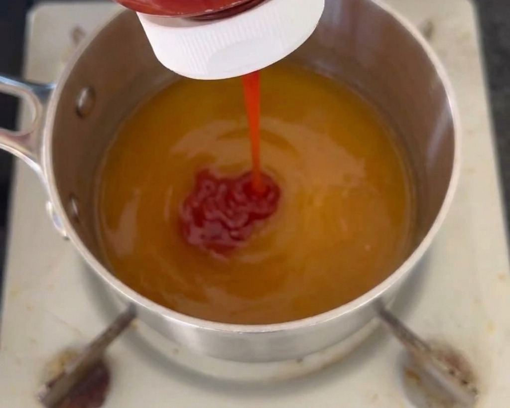 Un chorro de kétchup rojo saliendo de una botella hacia una olla que contiene una base líquida de salsa de color amarillo.